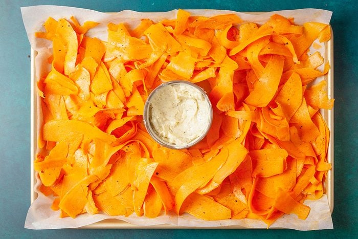 Overhead shot of a tray lined with parchment paper holding thin orange butternut squash ribbons arranged around a small bowl of creamy white dip in the center;