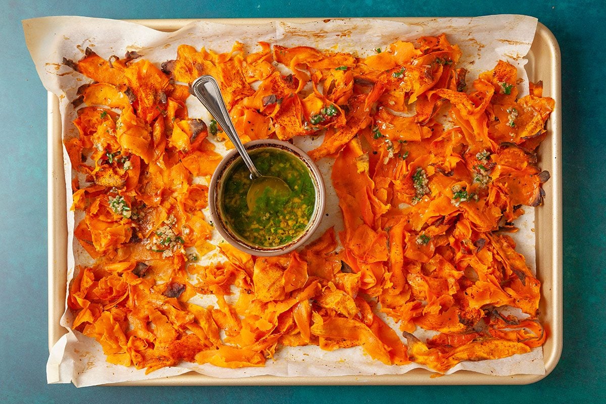 Overhead shot of a parchment-lined sheet pan holding roasted, thinly sliced sweet potatoes, crisped around the edges, with a small bowl of green herb sauce and a spoon placed in the center;