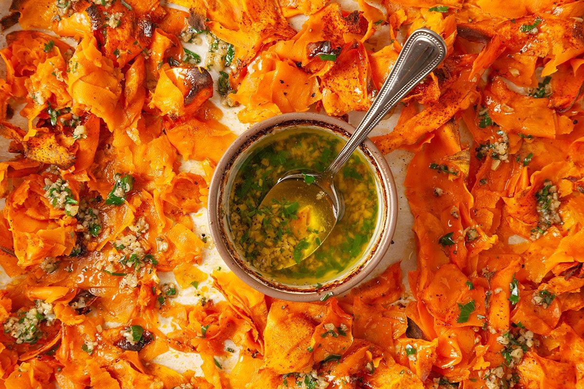Overhead shot of a bowl of green herb sauce with a spoon on a tray surrounded by roasted, thinly sliced sweet potatoes garnished with herbs and garlic;