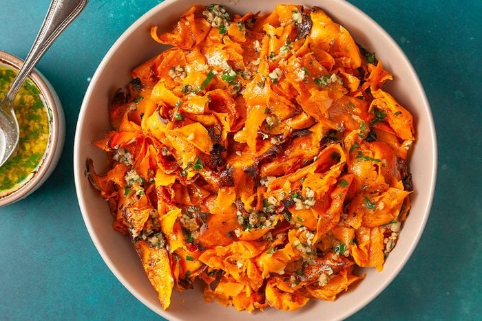 Overhead shot of a bowl filled with roasted sweet potato ribbons, lightly caramelized at the edges and tossed with a chunky green herb and garlic sauce; a small bowl of extra sauce sits nearby on a teal surface;