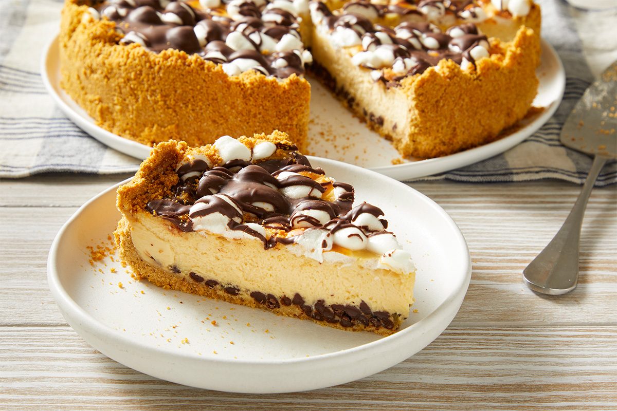 A slice of creamy pie with a graham cracker crust, topped with mini marshmallows and drizzled with chocolate, sits on a white plate. The rest of the pie is in the background on a wooden surface.