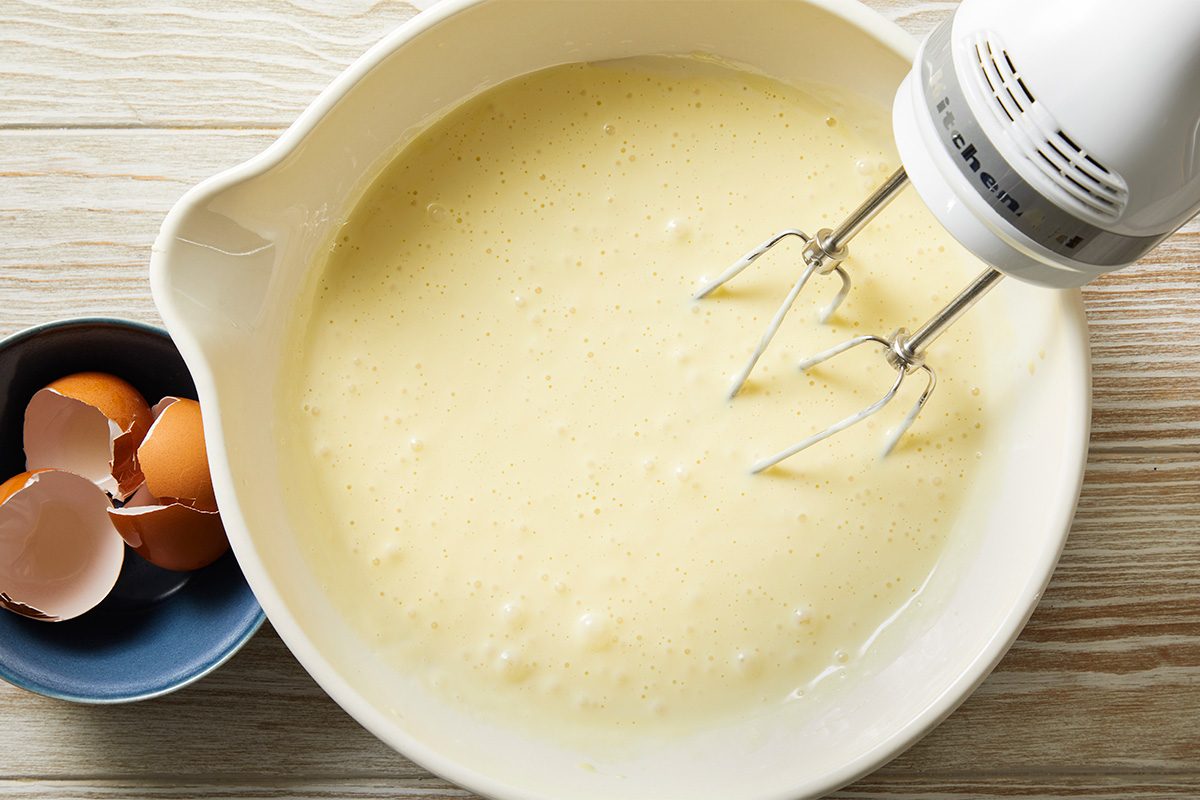 A mixing bowl filled with creamy batter and an electric hand mixer. Next to the bowl, a small dish holds empty eggshells, all on a light wooden surface.