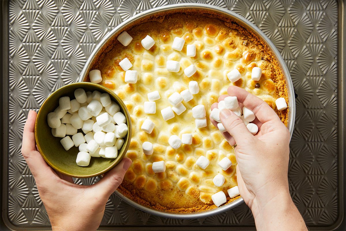 A person sprinkles mini marshmallows onto a round, baked dessert in a pan, with a bowl of marshmallows held in one hand, on a textured metal baking sheet.