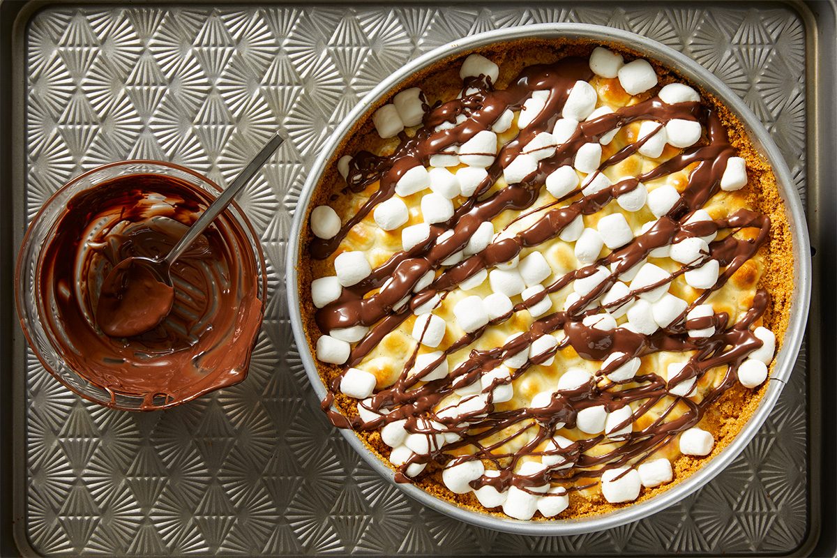 A round cake topped with mini marshmallows and drizzled chocolate sits on a patterned baking tray next to a bowl of melted chocolate with a spoon.