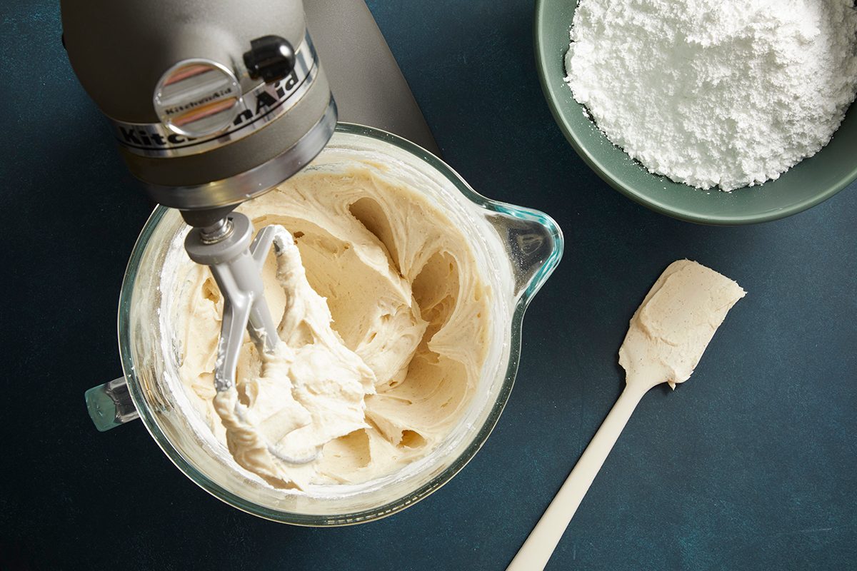 Overhead shot of a stand mixer bowl with whipped frosting, powdered sugar in a green bowl placed nearby.