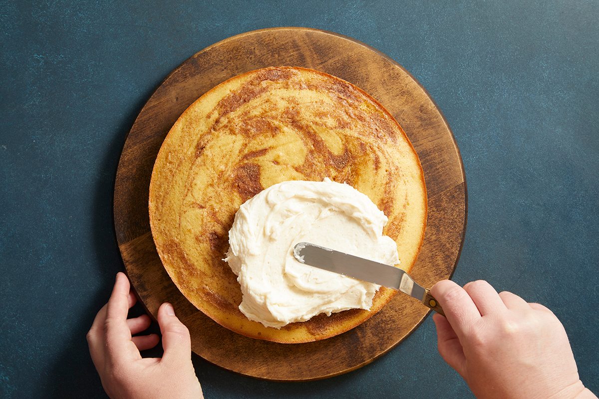 Overhead shot of a cake layer on a wooden turntable being frosted with a spatula, hand in frame spreading the icing.