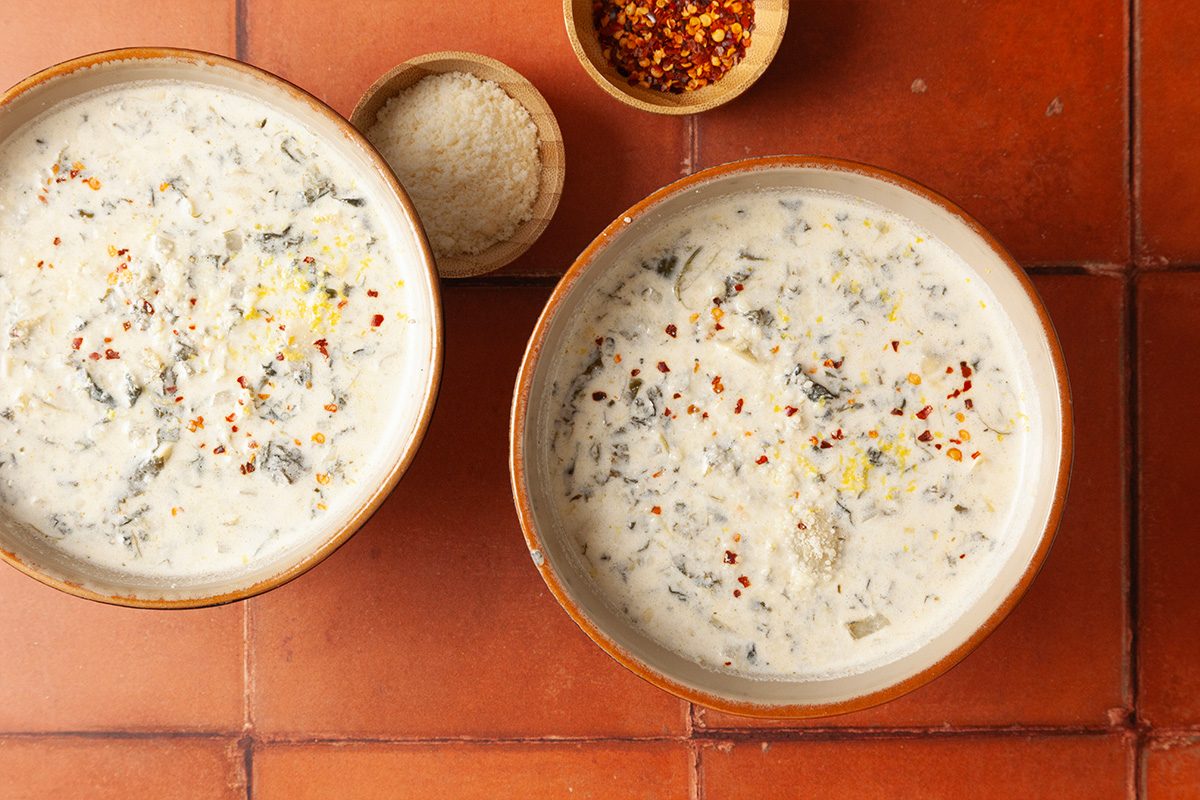 Overhead shot of two bowls of Spinach Artichoke Dip Soup garnished with red pepper flakes and lemon zest on terracotta tiles, with cheese and spices nearby;