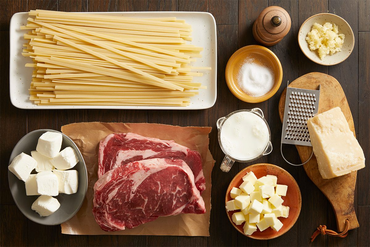 Overhead shot of raw ingredients for Steak Alfredo, uncooked pasta, ribeye steaks, garlic, cream, butter, and Parmesan arranged neatly on a wooden surface.