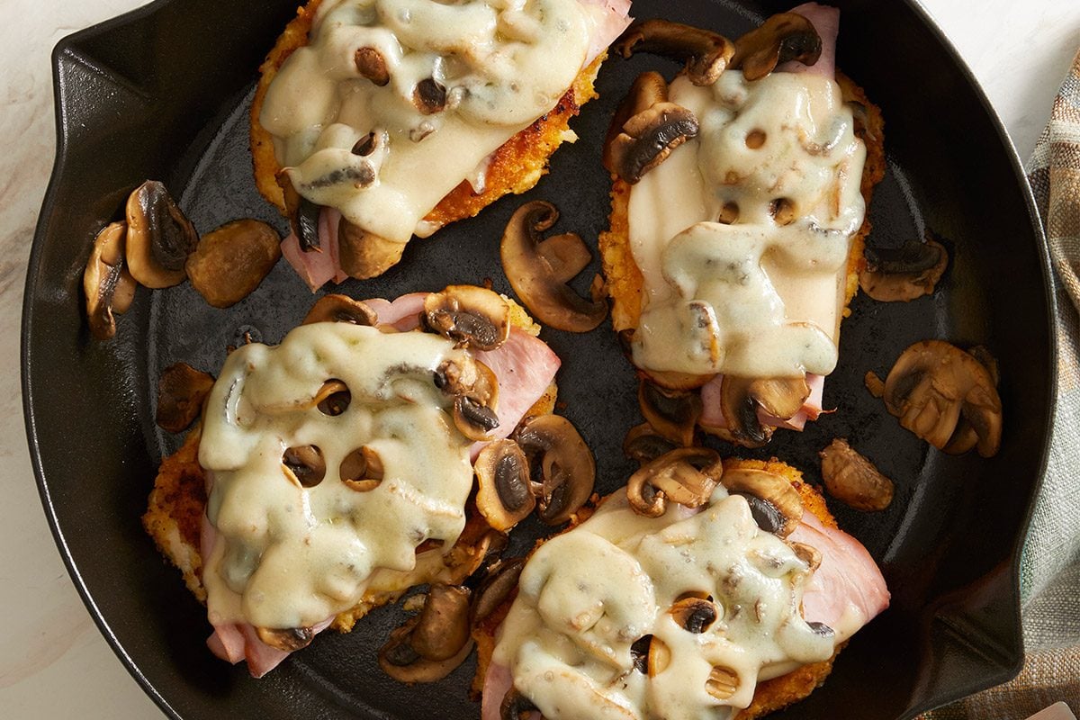 Vertical shot of Swiss Mushroom Chicken topped with sautéed mushrooms, ham, and melted Swiss cheese in a black skillet on a plaid towel.