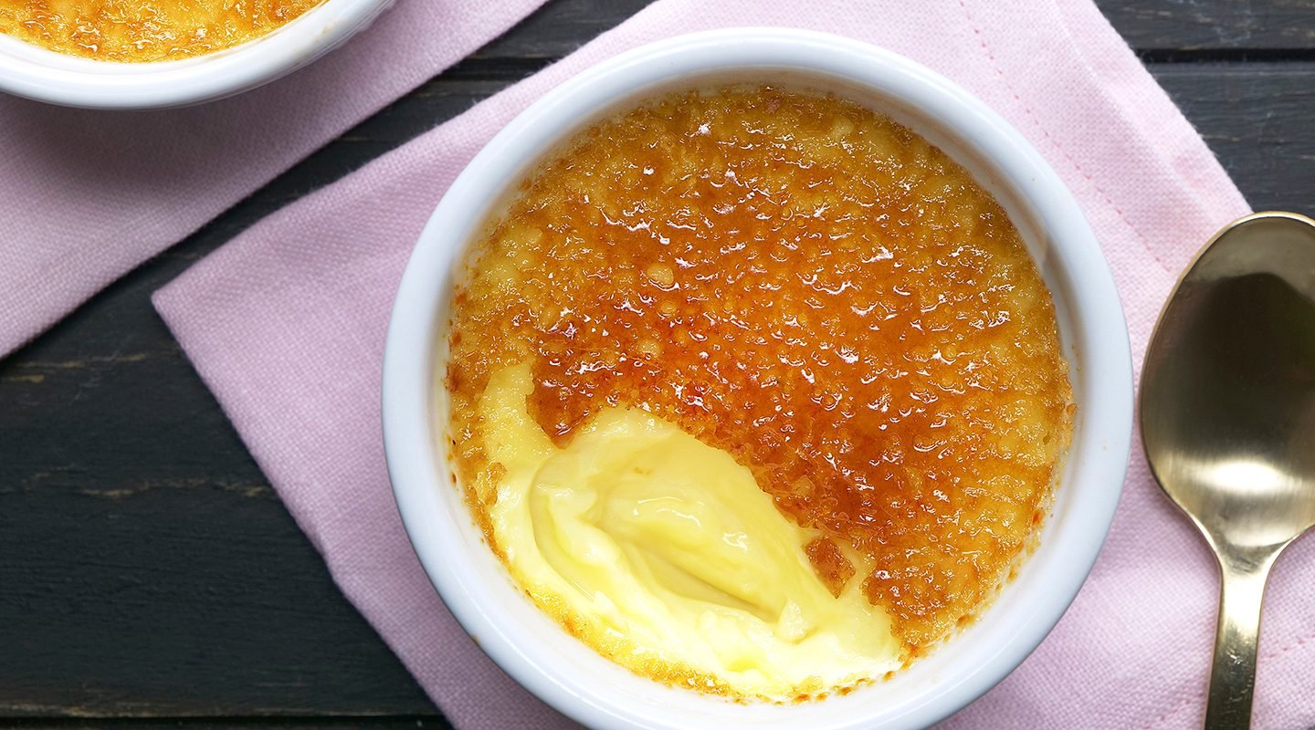 A ramekin of crème brûlée with a caramelized sugar crust, partially cracked to reveal creamy custard underneath, sits on a pink napkin next to a gold spoon.