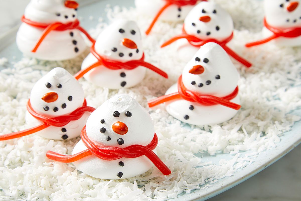 3/4th shot of 3D Meringue Snowmen with candy faces and red licorice scarves They sit on a plate of shredded coconut that looks like snow