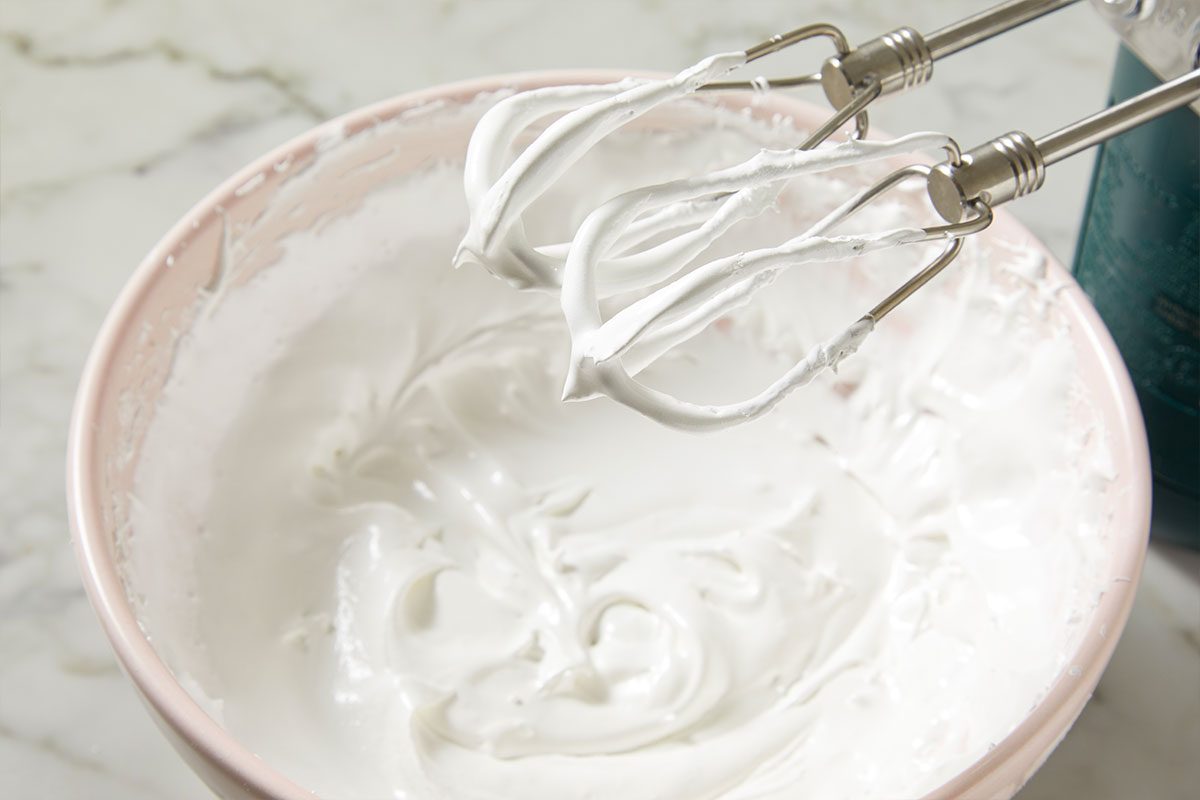 3/4th shot of a close-up shows a mixing bowl with glossy stiff meringue The metal beaters from a hand mixer rest above the mixture on marble