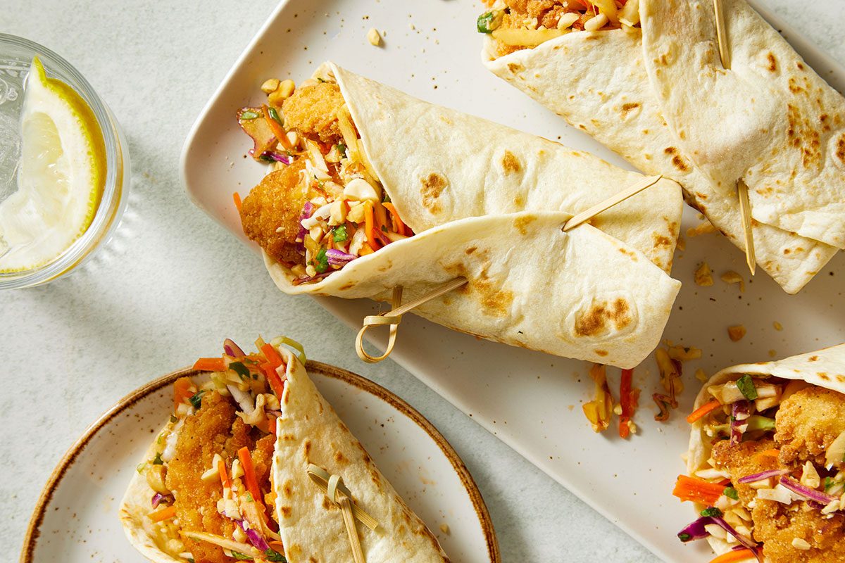 Overhead view of Asian Chicken Crunch Wraps on a white plate with visible crispy chicken and slaw filling, accompanied by a small bowl of sauce and a few wrap pieces scattered nearby.