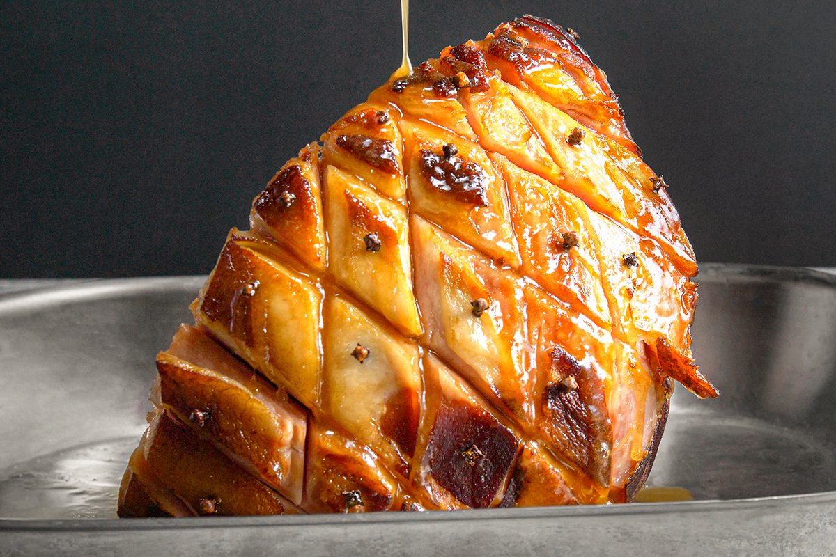 A glazed ham is scored in a diamond pattern and topped with whole cloves, with a golden drizzle being poured over it, set in a metal roasting pan.