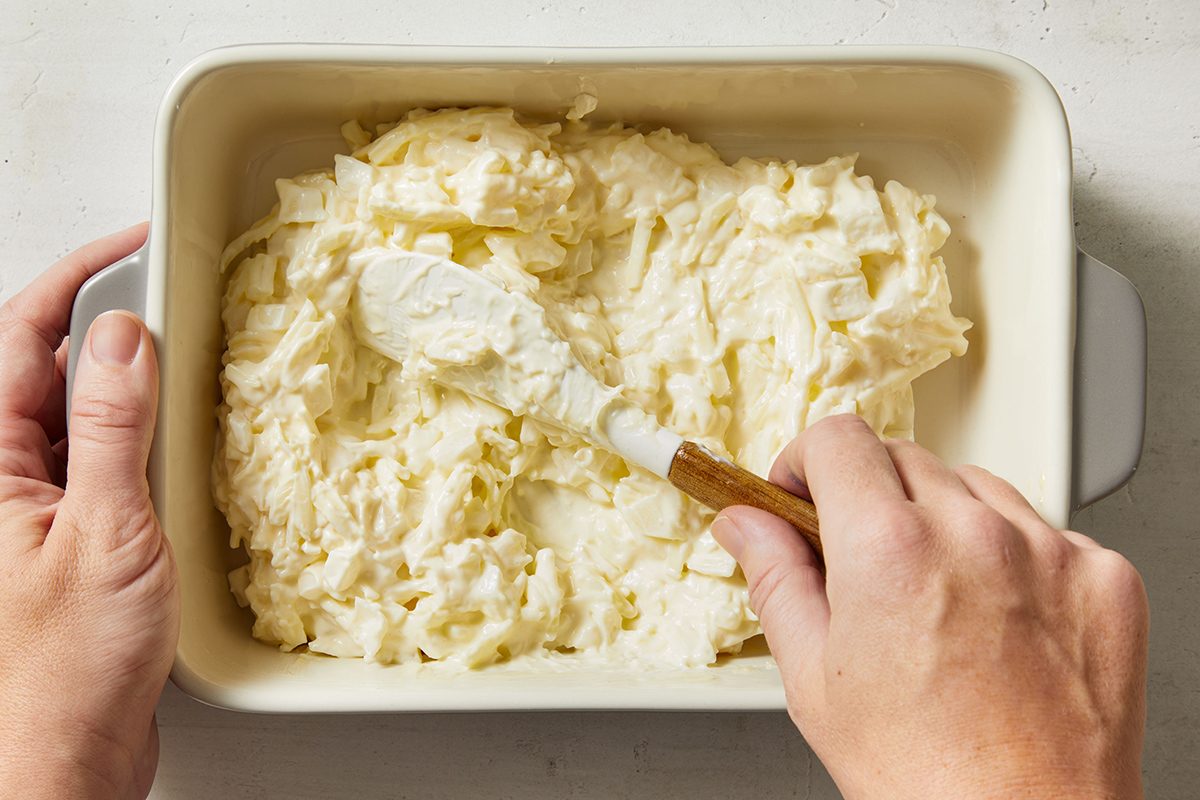 A person spreads a creamy, shredded mixture evenly in a white rectangular baking dish using a spatula.