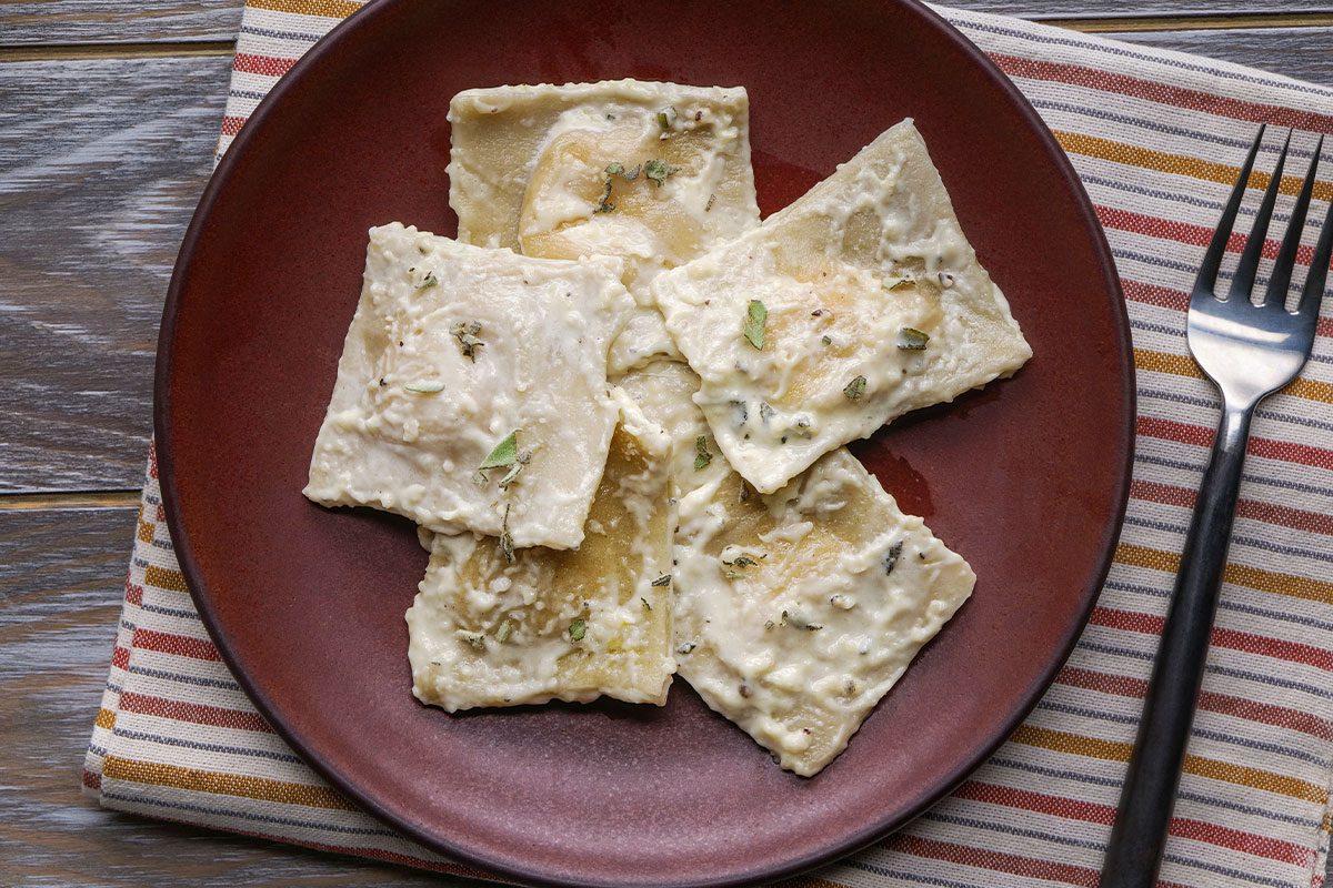Overhead shot of Butternut Squash Ravioli covered in creamy white sauce and herbs rests on a striped cloth napkin with a fork to the right of the plate