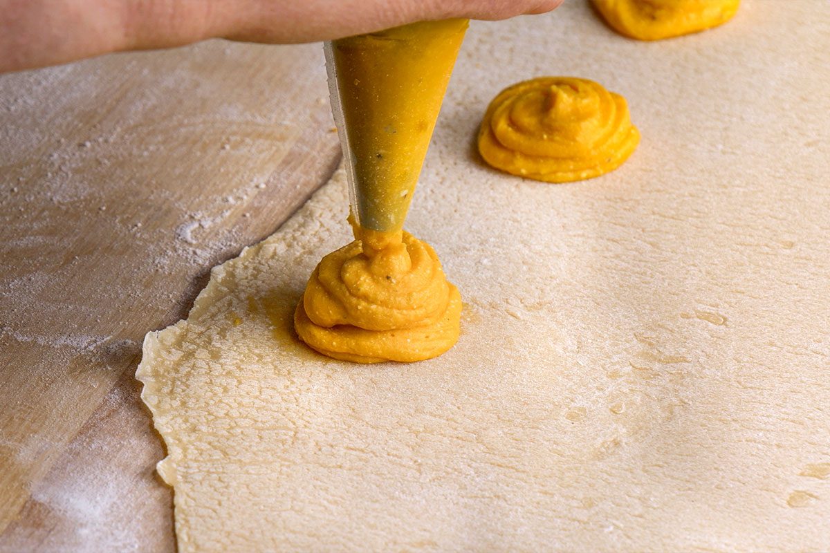Closeup shot of a hand is holding a piping bag and carefully squeezing orange filling onto pasta dough the dough rests on a wooden surface