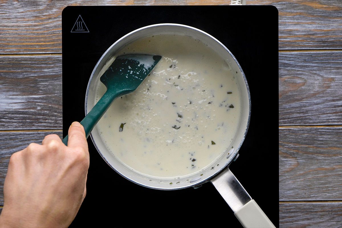 Overhead shot of a hand stirs a creamy white sauce with herbs in a saucepan using a green spatula on a black stovetop and the sauce looks smooth It is gently mixed
