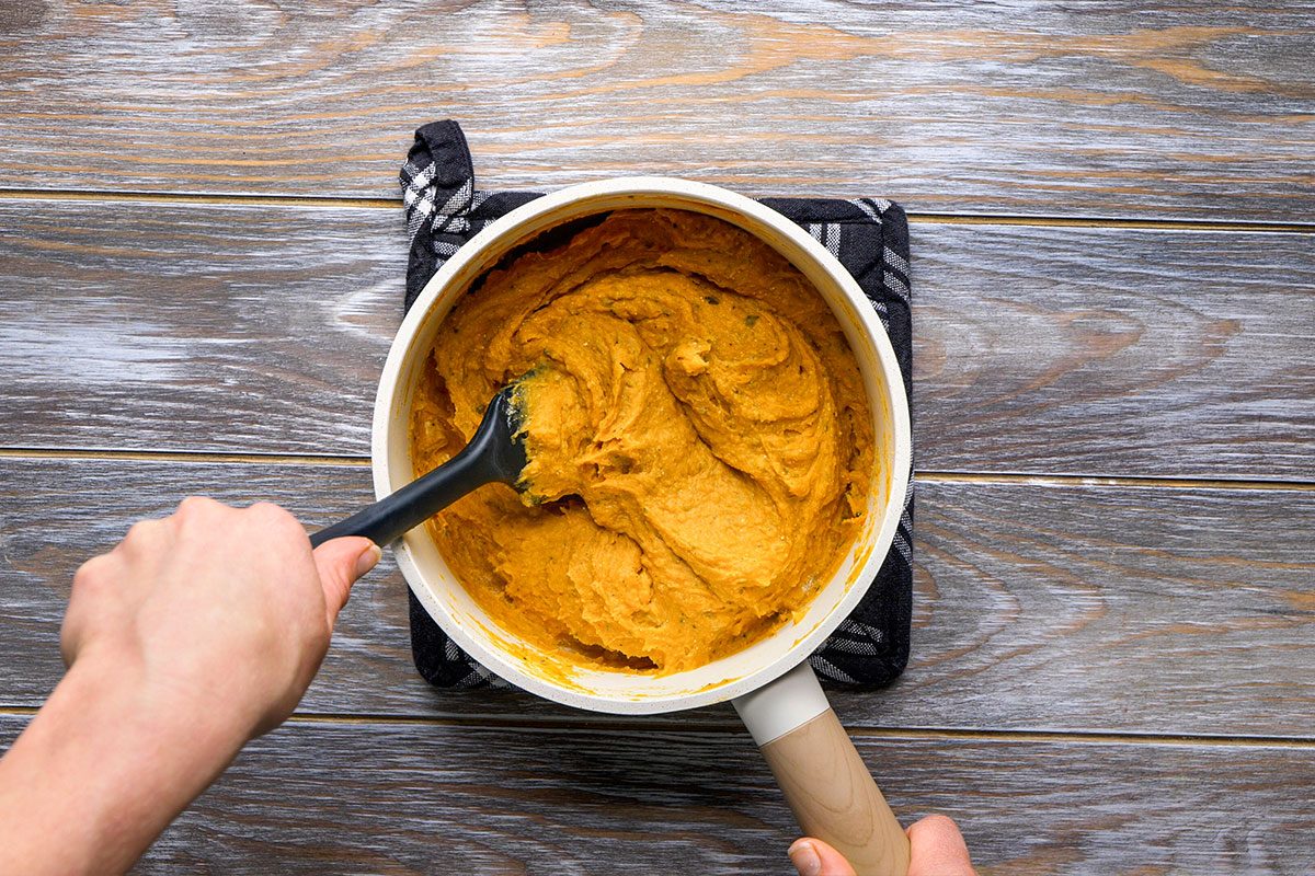 Overhead shot of a person stirs thick orange mashed butternut squash in a white saucepan using a black spatula and the pot sits on a dark potholder on a wooden surface