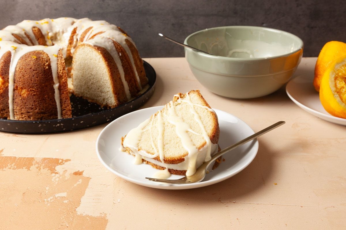 3/4th Shot of a slice of Cardamom Pound Cake with white icing is on a plate with a fork nearby the rest of the cake and bowl of icing and a halved orange