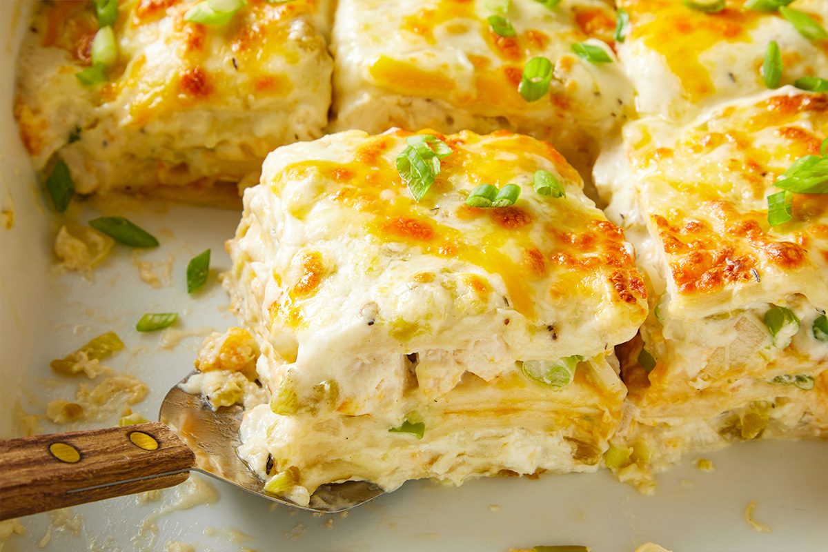 Close-up shot of a square piece of Chicken Chile Lasagna being lifted from the pan, showing creamy layers of tortillas, chicken, sauce, and melted cheese.