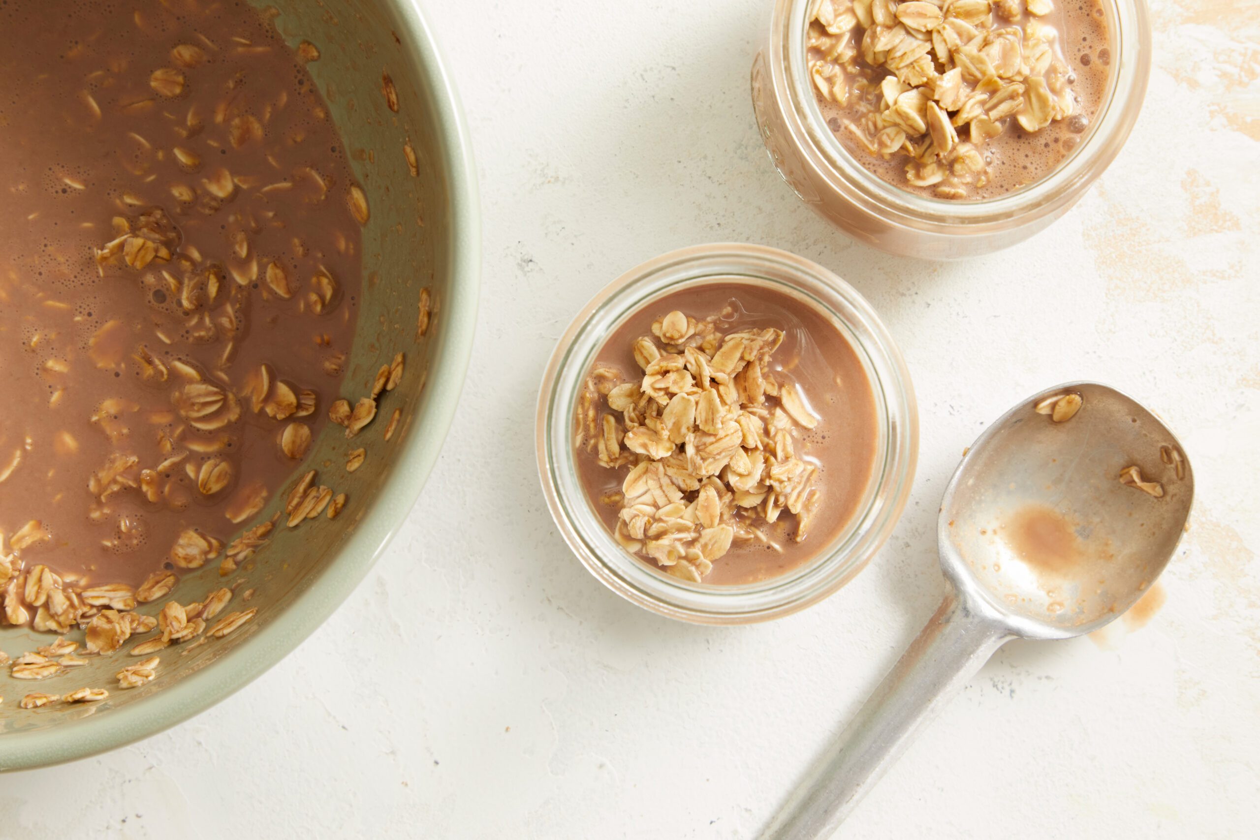 Overhead shot of a large bowl of chocolate oat mixture with two smaller jars being filled beside it, a spoon resting nearby.