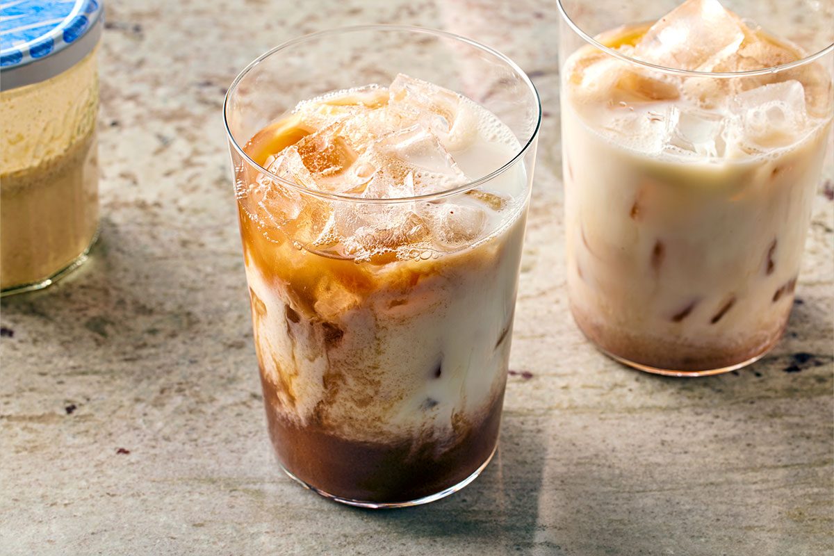 3/4 angle view shot of two clear glasses filled with iced coffee and milk, showing swirling layers of brown coffee and white milk, with ice cubes floating on top, placed on a light stone surface;
