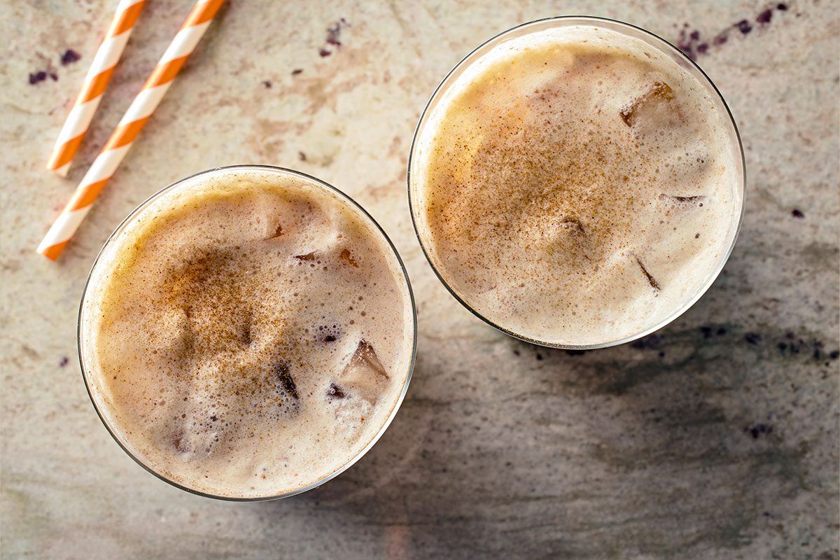 Overhead shot of two Iced Pumpkin Cream Chai Tea Lattes topped with foam and sprinkled with cinnamon, each with a striped straw, on a light surface;