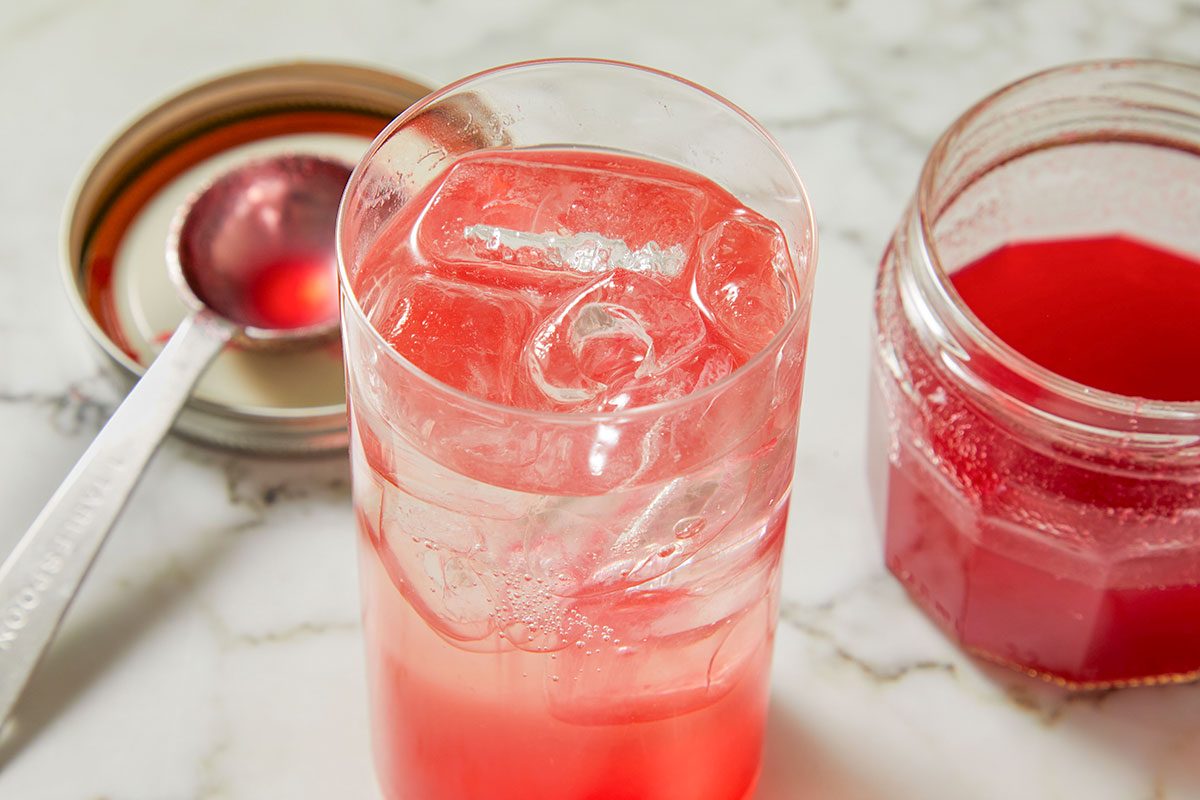 Overhead shot of a tall glass filled with ice and pale pink ginger ale, with a jar of cranberry syrup and ladle beside it.