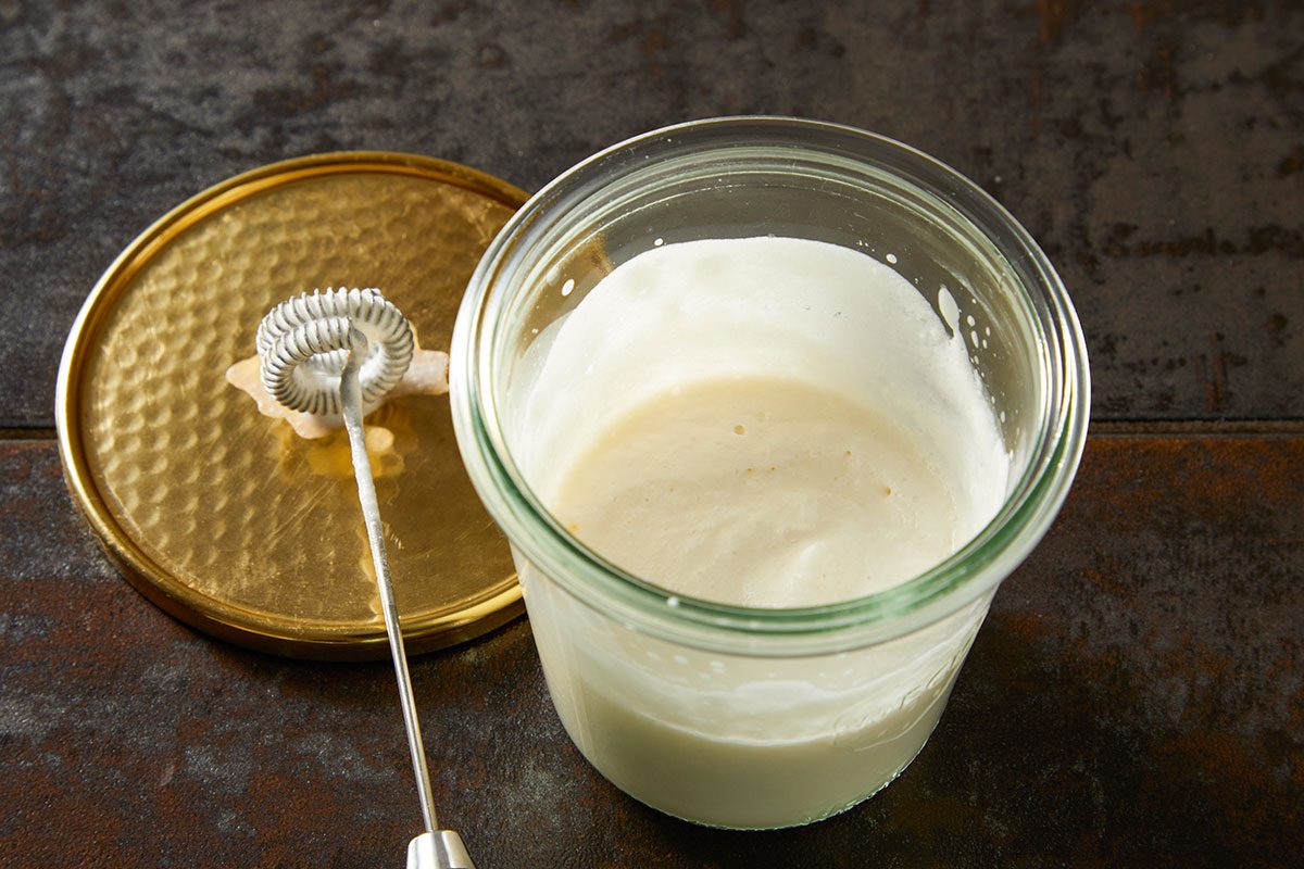 Closeup shot of creamy yogurt mixture in a glass jar with a whisk, set on a dark textured surface with a gold lid nearby;