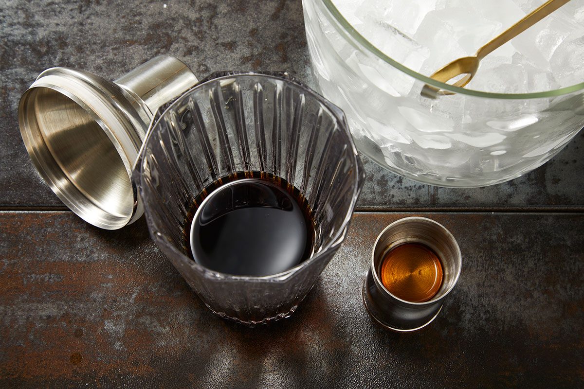 Overhead shot of cocktail ingredients including a mixing glass with dark liquid, ice bowl with spoon, and metal jigger arranged on a dark textured surface;