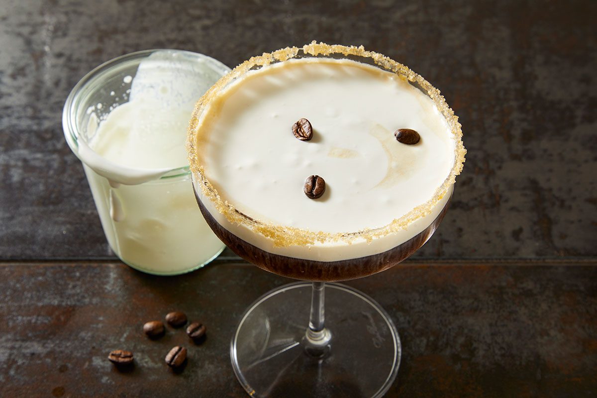 3/4 angle close-up shot of a creamy coffee cocktail in a coupe glass with a sugared rim, garnished with coffee beans, set on a dark textured surface;
