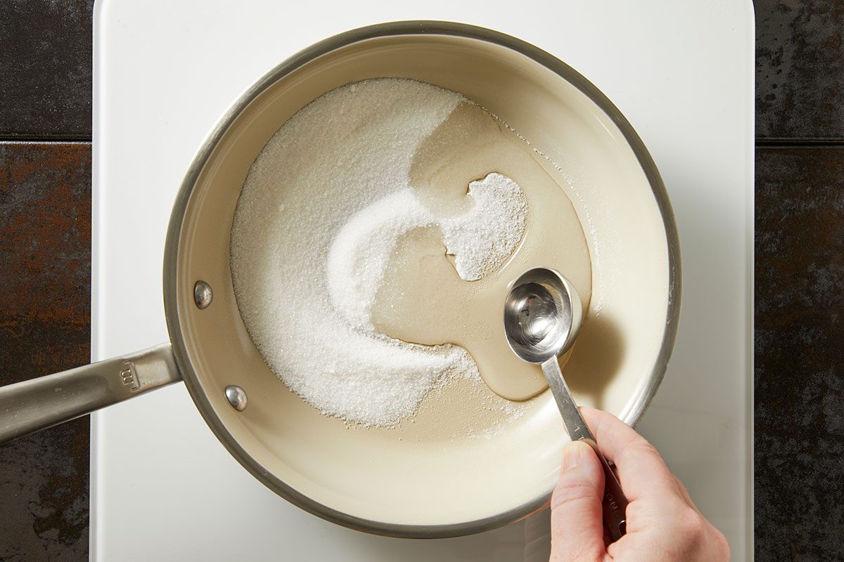 Overhead shot of sugar and liquid being combined in a saucepan, with a measuring spoon stirring on a stovetop;