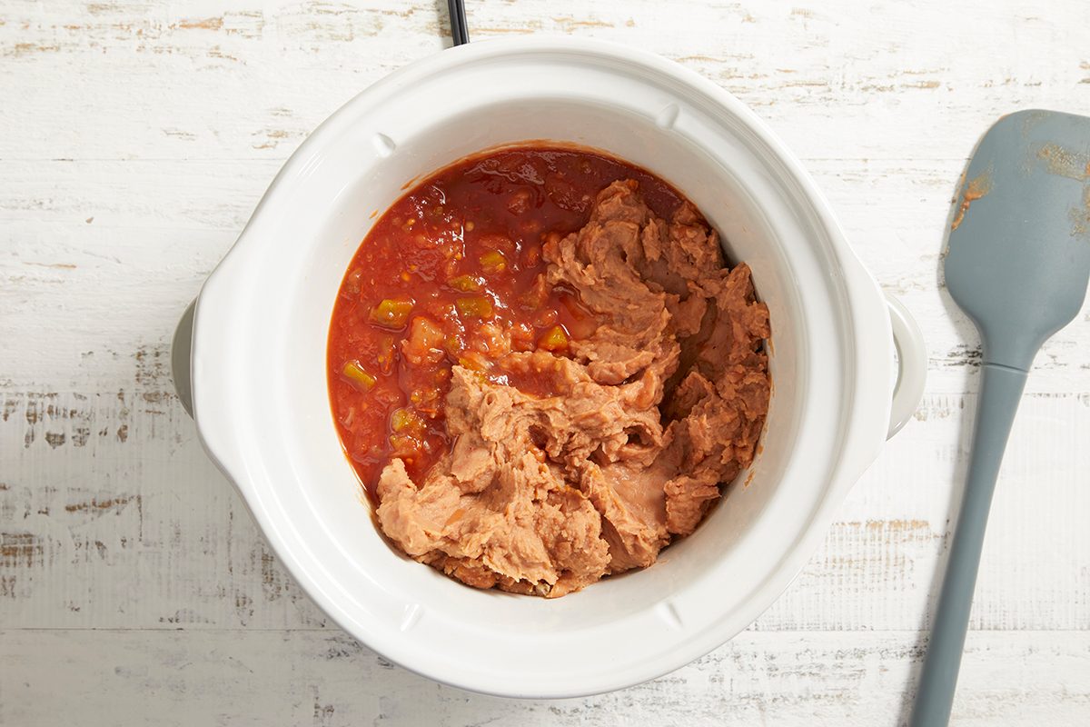 A white slow cooker filled with canned refried beans and tomato salsa sits on a white wooden surface next to a gray spatula.