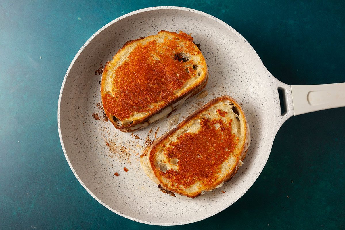 Overhead shot of two assembled sandwiches toasting in a light-colored skillet until golden brown; The bread is evenly crisped with a buttery surface, set against a teal background;