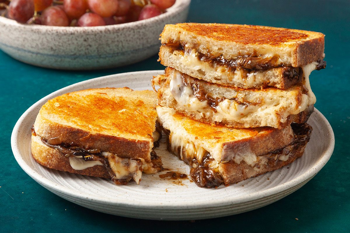 Close shot of French onion grilled cheese sandwiches arranged on a light ceramic plate; One sandwich is cut to reveal layers of caramelized onions and melted cheese, while the remaining halves are stacked for height; A bowl of grapes softly blurs in the background, adding a warm, inviting finish;