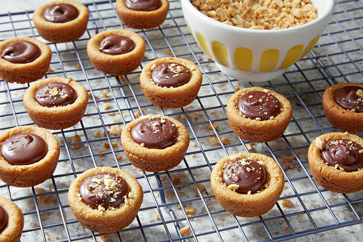 Peanut butter cookie cups filled with chocolate and sprinkled with crushed peanuts rest on a cooling rack next to a bowl of chopped peanuts.