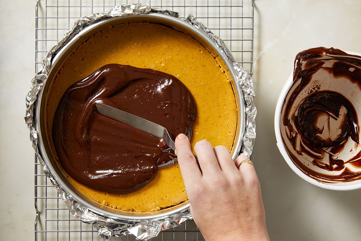 Overhead shot of glossy chocolate ganache being spread over the cooled cheesecake with a spatula, coating the top evenly.