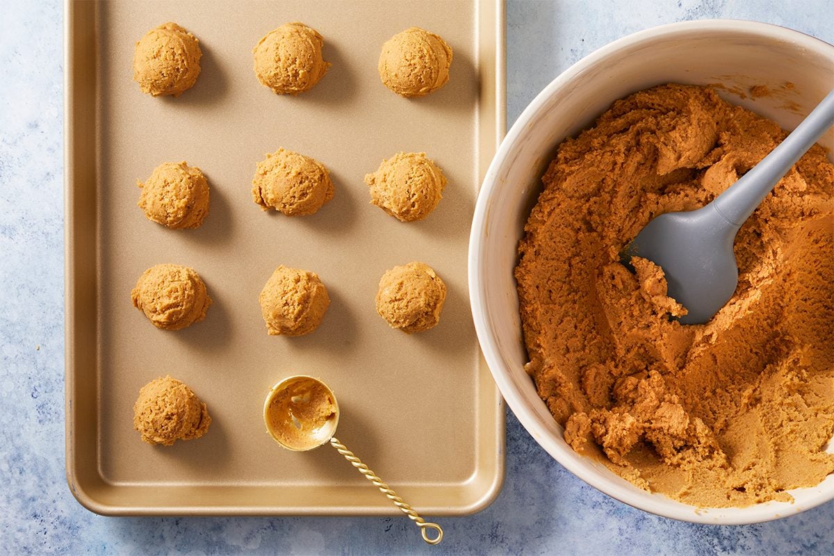 Overhead shot of a baking sheet has twelve scoops of cookie dough and it rests near a bowl filled with more dough with a gray spatula and gold scoop both are placed on a light blue surface