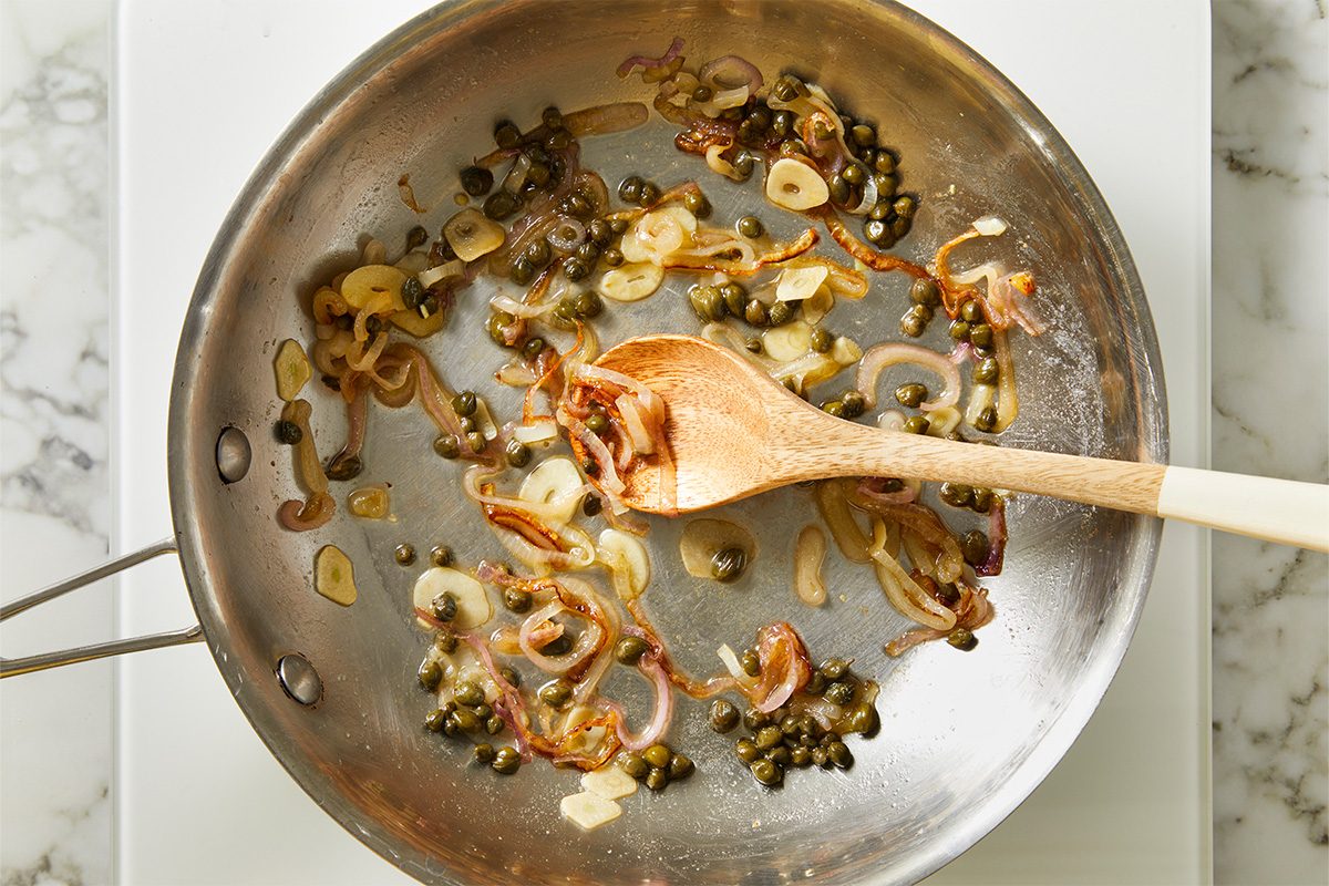 Overhead shot of sliced shallots, garlic, and capers sautéing in a skillet, being stirred with a wooden spoon on a stovetop;