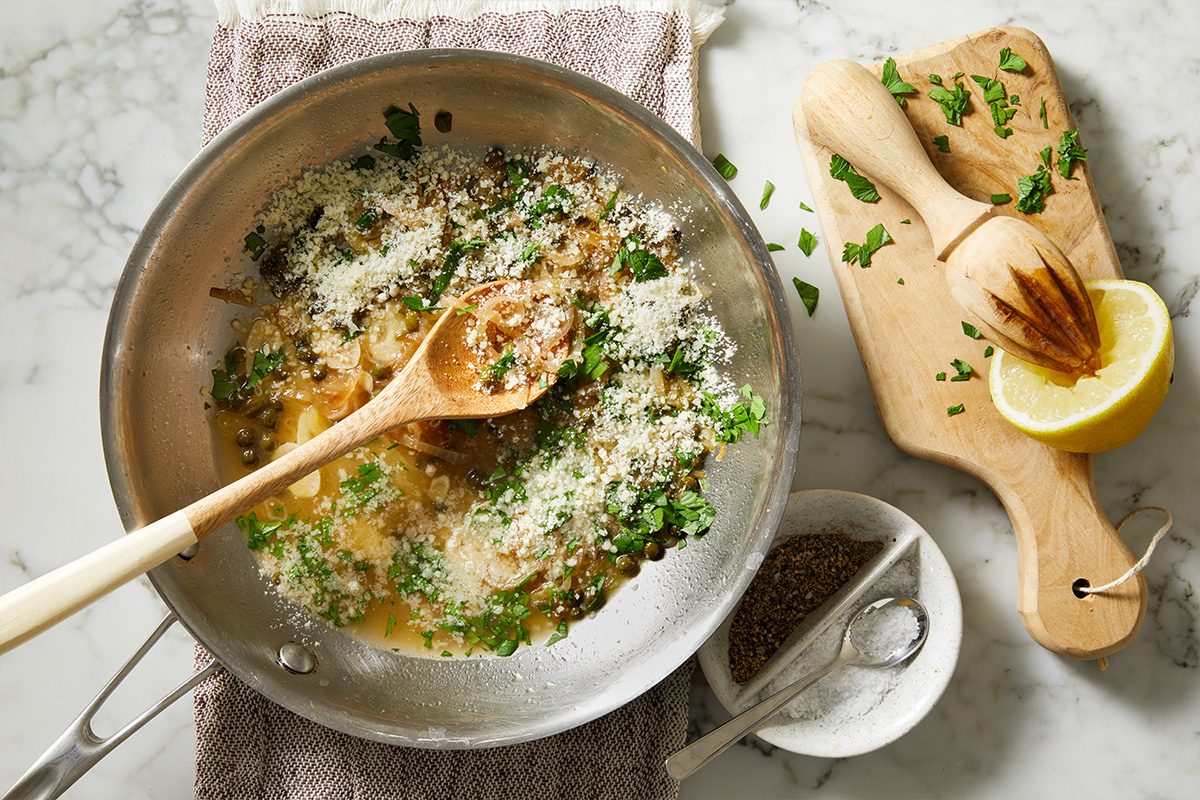 Overhead shot of a skillet with a caper and shallot sauce finished with grated cheese and fresh herbs, with lemon and seasoning tools alongside on a light marble surface;