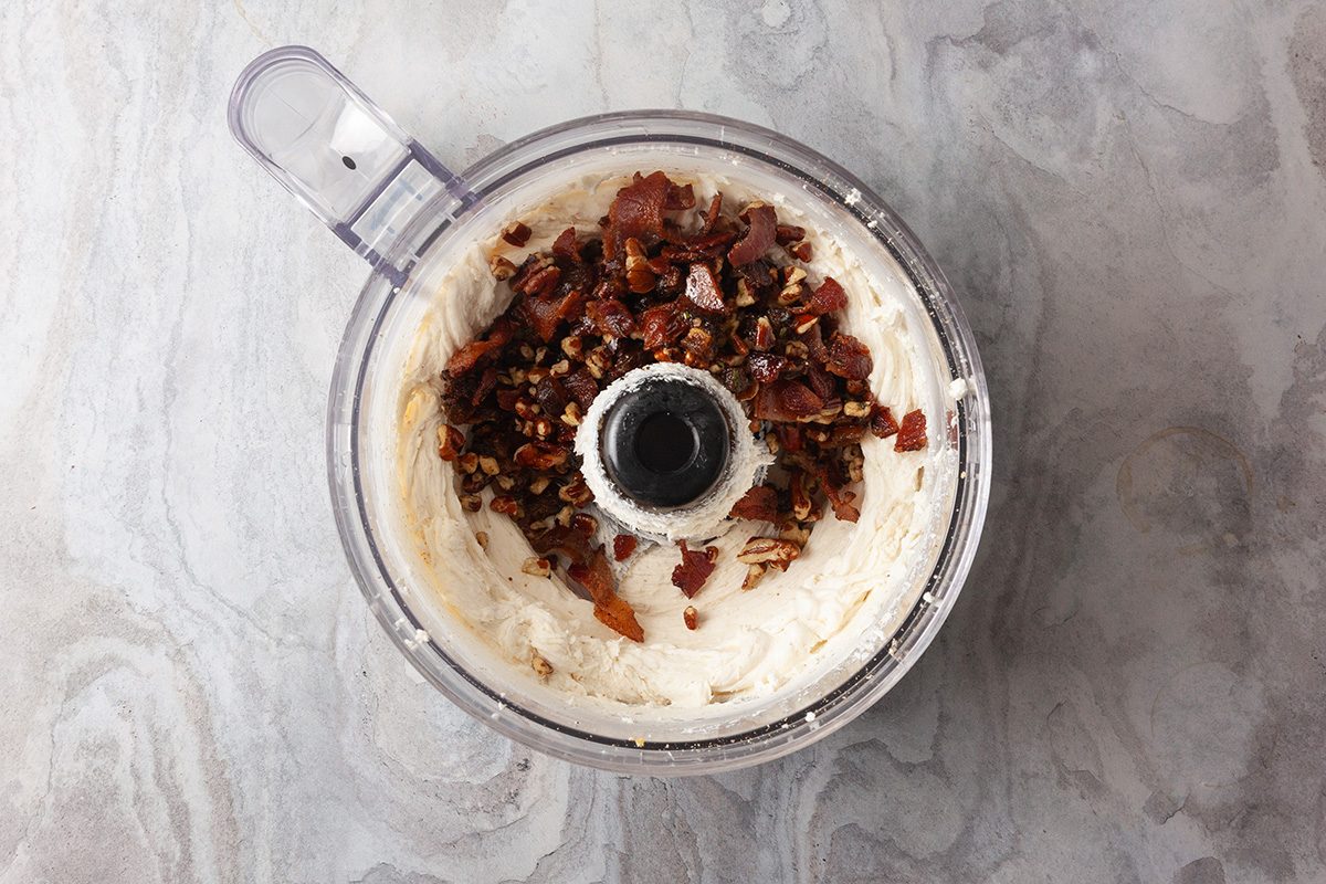 Overhead shot of a food processor holds creamy white cheese mixture and is topped with chopped pecans and crumbled bacon on a light gray marble surface