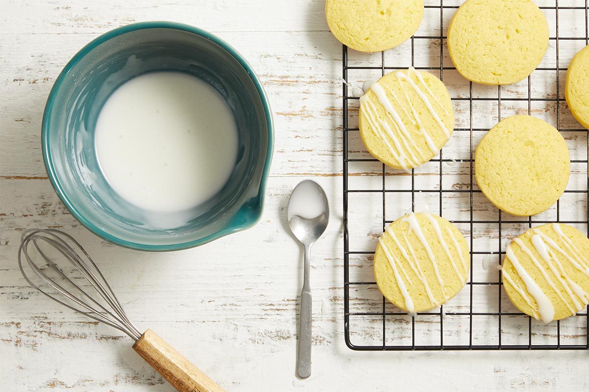 Overhead view of baked lemon pudding cookies cooling on a wire rack with a bowl of glaze and whisk nearby.