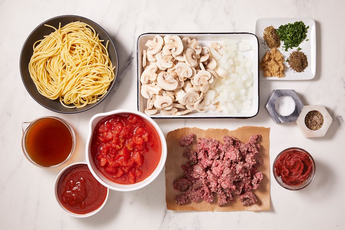 Various ingredients for making spaghetti are arranged on a white surface, including uncooked pasta, ground beef, sliced mushrooms, chopped onions, canned tomatoes, tomato paste, broth, and assorted seasonings.