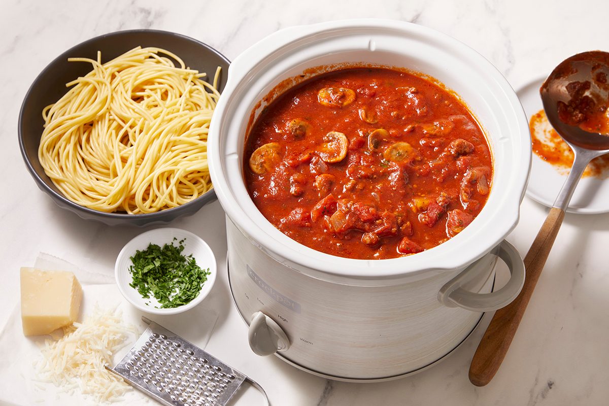 A white pot filled with tomato-based pasta sauce with mushrooms sits next to a bowl of cooked spaghetti, a bowl of chopped parsley, a block of cheese, and grated cheese on a marble surface.