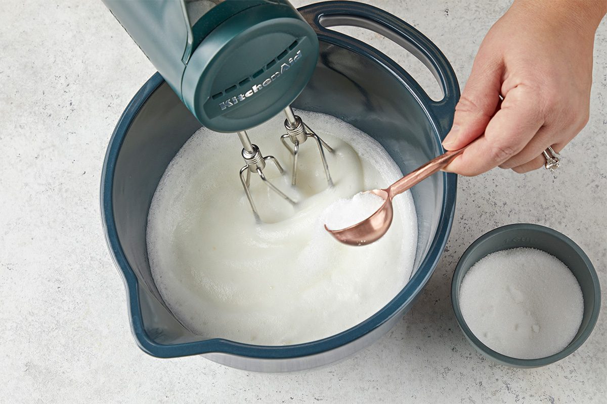 Overhead shot of a person adding sugar from a spoon into a mixing bowl of whipped egg whites while using an electric hand mixer, with a small bowl of sugar nearby on the counter;