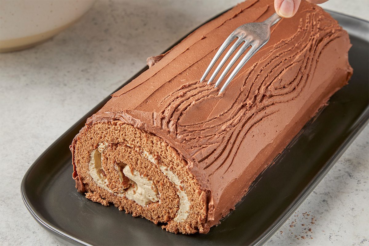 Closeup shot of a person using a fork to create wavy lines in the chocolate frosting of a swirled cake roll with cream filling, placed on a black rectangular tray;