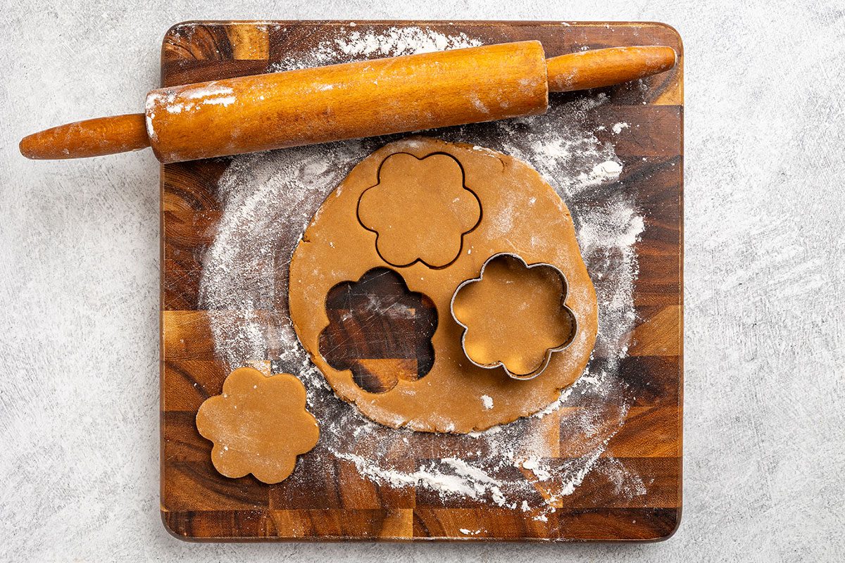 Overhead shot of a wooden cutting board with rolled-out dough; a rolling pin, and flower-shaped cookie cutters making cutouts; surrounded by scattered flour.