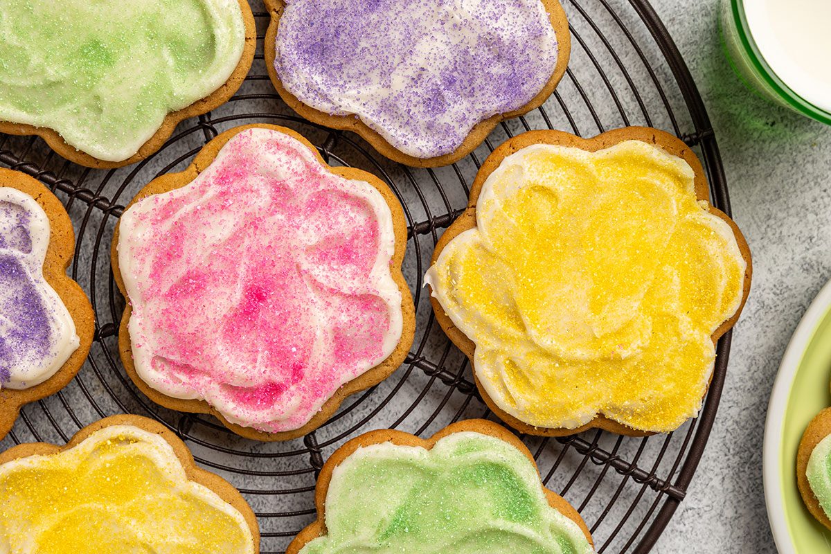 Overhead shot of Peanut Butter Cutout Cookies shaped like flowers; decorated with pastel-colored frosting in pink, yellow, green, and purple; Some cookies are arranged on a cooling rack; with glasses of milk nearby; all set on a light textured surface.