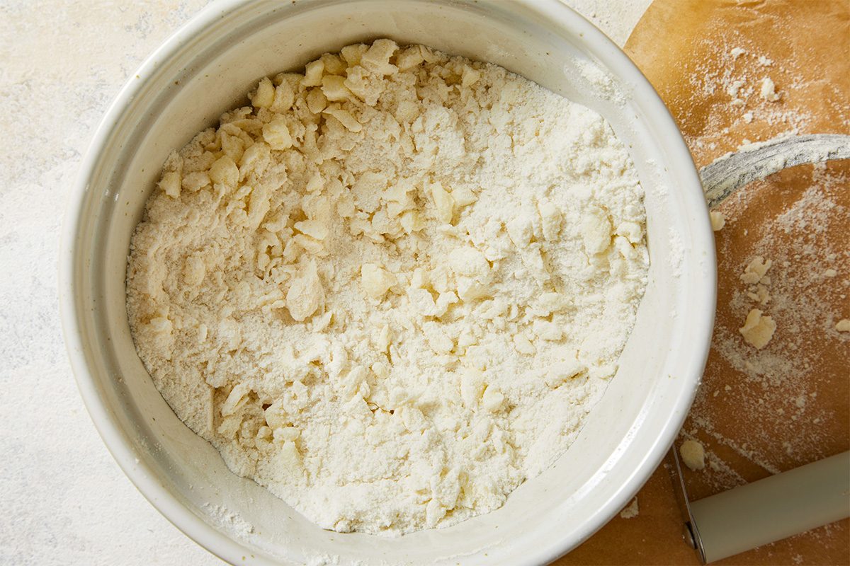 Overhead shot of flour and butter mixture in a white mixing bowl on a wooden surface, showing crumbly dough texture.